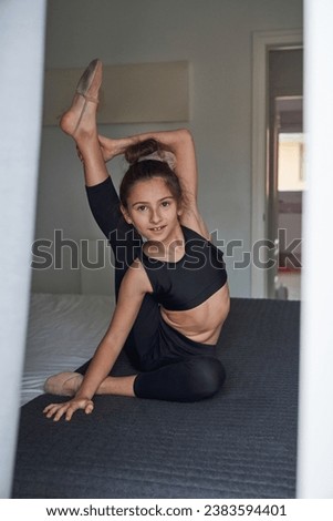 Full body of fit preteen girl in black top and leggings sitting on bed in bedroom and stretching leg up in vertical split while looking at camera during gymnastics training