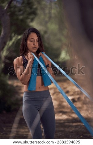 Strong ethnic lady in sportswear performing intense exercises with elastic band while standing in green woods