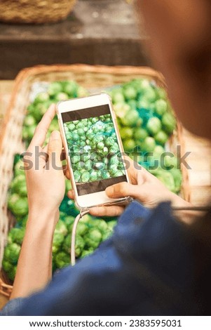 From above of crop unrecognizable person with mobile phone taking picture of artichoke pile in wicker basket in market