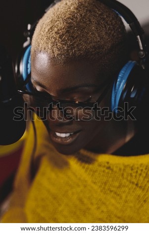 From above of positive young african american female singer with short hair wearing eyeglasses and headphones while recording song via microphone in studio