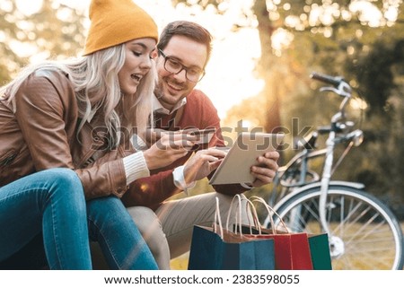 Young couple shopping online with credit card on digital tablet pc in the city park. Two friends booking a vacation together while resting in city park after shopping.