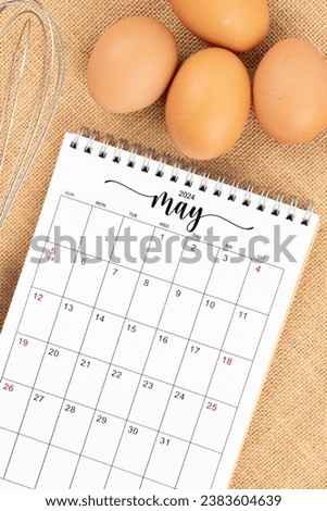 May 2024 Desk calendar and fresh eggs with egg whisk on burlap background.