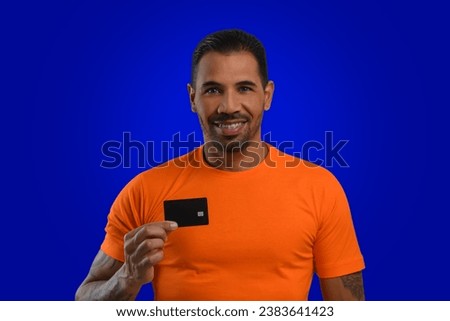 A man with a black credit card in his hand, wearing an orange T-shirt, looks at the camera and smiles, blue background