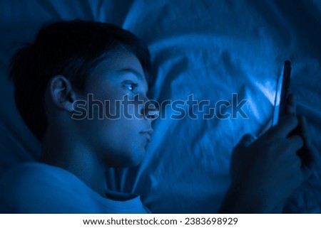Boy using a smartphone at night in bed. 10 year old boy playing with mobile phone before going to sleep. Concept of children using the smartphone in their room alone without control. Copy space