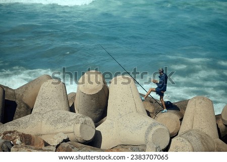 Rear view of unrecognizable fisherman holding fishing rod, standing on the seashore on breakwater and fishing in Atlantic Ocean. People. Leisure. Hobby Royalty-Free Stock Photo #2383707065