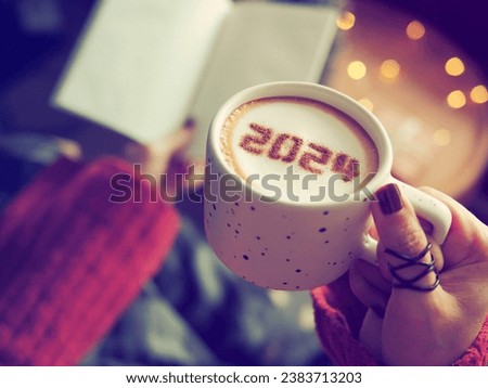 New year 2024 celebrated coffee cup with number 2024 over frothy surface holding by female hands with nail polish and rings, blurred background with a book, wooden round table, Christmas string lights