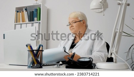 Double checking patients histories done by nurse using computer and paper prints. Elderly female doctor writing perscriptions and notes them on clipboard and laptop.