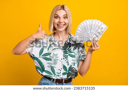 Photo portrait of attractive young woman thumb up hold money fan dressed stylish leaves print clothes isolated on yellow color background