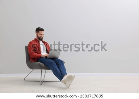 Handsome man using laptop while sitting in armchair near light grey wall indoors, space for text