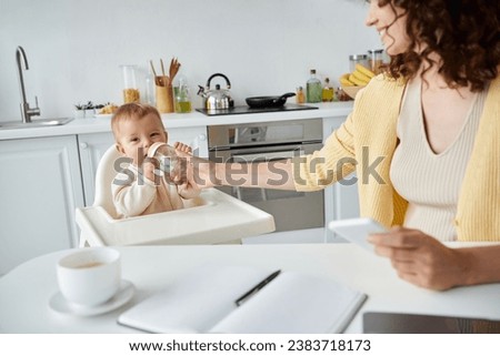 woman with mobile phone helping little girl drinking from baby bottle while working in kitchen