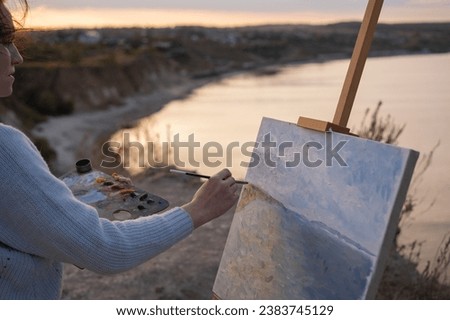 A young woman stands on a beach at sunset and paints an oil painting on an easel. The artist paints a picture of the river bank.