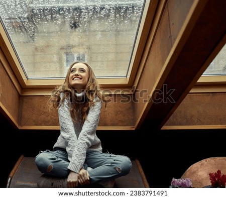 Young girl is happy about the rain outside the window, sitting in her cozy room under the window in the ceiling, on which the rain flows. Concept of inner balance, joy, good news, romantic feelings