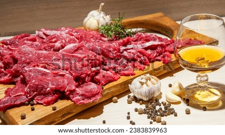 A wooden cutting board with red cube meat decorated with garlic and spices next to a glass of wine