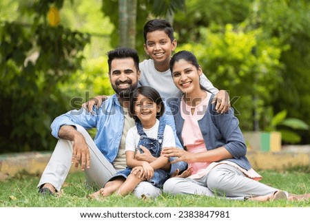 Happy indian couple with kids embracing by looking at camera while sitting at park - concept of relationship, family time and togetherness Royalty-Free Stock Photo #2383847981