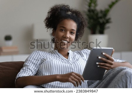 Head shot portrait of happy relaxed millennial Black girl, young woman resting on couch with gadget, using tablet computer, reading book, watching movie, working from home, smiling at camera