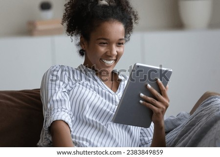 Happy excited millennial girl, young Black woman reading book on tablet computer, watching funny movie, making video call, talking at pad webcam, relaxing on sofa at home, smiling, laughing