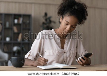 Serious African American millennial student watching learning webinar, virtual training, online workshop on smartphone, writing notes in notebook, studying from home, taking remote course Royalty-Free Stock Photo #2383849869