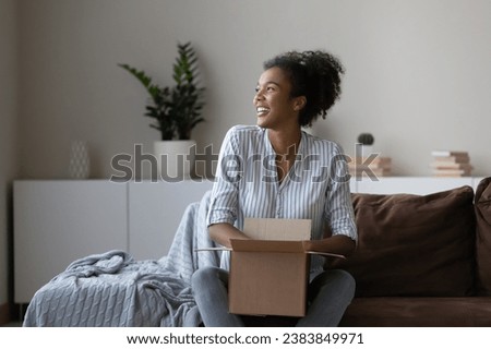 Happy young African woman, millennial shopper, online store customer opening carton box at home, unpacking delivery parcel received from internet shop. Unboxing package, smiling. Shipping concept