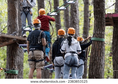 A group embarks on a high ropes challenge at an adventure park, showcasing teamwork and agility amid treetop obstacles. Royalty-Free Stock Photo #2383853125