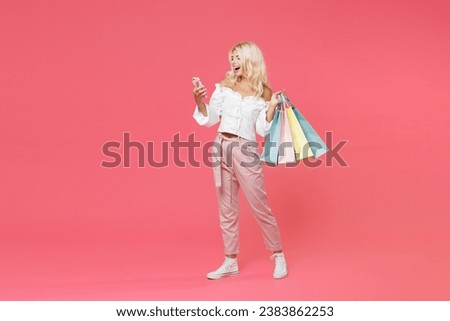 Full length portrait of funny young blonde woman 20s in white casual clothes hold package bag with purchases after shopping using mobile cell phone isolated on bright pink colour background studio