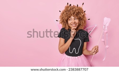 Positive curly haired woman pretends being tooth fairy poses with magic wand dressed in costume comes on carnival focused aside happily isolated over pink background copy space for your promotion Royalty-Free Stock Photo #2383887707