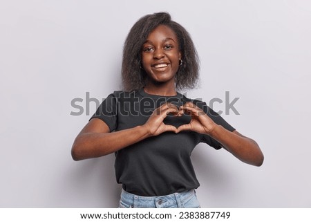 Horizontal shot of pretty dark skinned woman shows heart sign I love you gesture smiles happily dressed in casual black t shirt and jeans isolated over white background. Romantic feelings concept