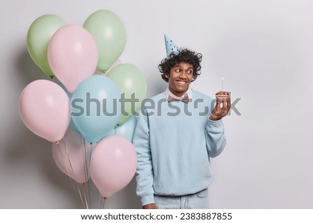 Indoor shot of cheerful Hindu man looks at small cupcake with glad expression wears party cone hat and jumper with bowtie comes on party to celebrate something stands near bunch of inflated balloons Royalty-Free Stock Photo #2383887855