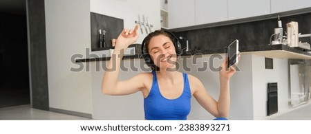 Cheerful brunette girl does fitness at home, wears wireless headphones and dances, holds smartphone, does workout indoors on floor, wears blue activewear.