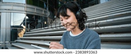 Young woman draws on her tablet, listens to music in headphones. Asian girl sits on stairs and does graphic design project, sits on street stairs.