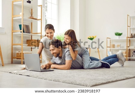 Happy young parents with their child girl lying on the floor at home using laptop. Smiling family enjoying weekend watching funny cartoon online or talking on video call. Family leisure concept.