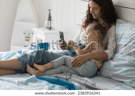 Young loving mother relaxing in bed hug her little cute daughter, spends family weekend at home together, use mobile phone to watch videos, shopping online, enjoy new applications. Leisure on internet
