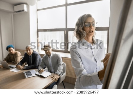Serious pretty mature female business leader writing on whiteboard on meeting with multiethnic team, explaining project plan, strategy, sharing ideas with office colleagues, giving report, seminar Royalty-Free Stock Photo #2383926497