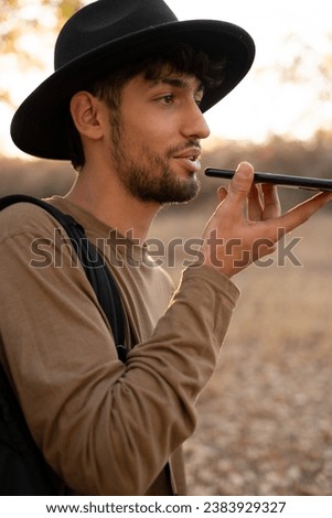Male traveler wearing hat using a smartphone voice recognition online, walking and talking outdoor. Voice chat bot, conversational AI. Copy space