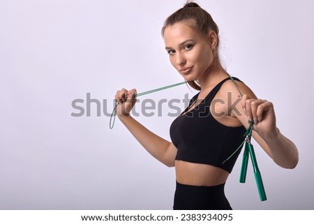 Young beautiful fitness trainer girl performing exercise with skipping rope in gym. On a gray background, a slender athletic female athlete poses sideways with a skipping rope in her hand