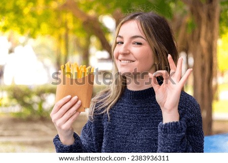 Young pretty Romanian woman holding fried chips at outdoors showing ok sign with fingers
