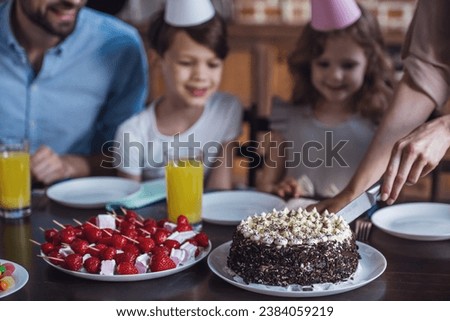 Happy family is sitting at the table in decorated kitchen during birthday celebration. Cropped image of mom cutting birthday cake