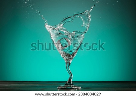 Dynamic action of ice cube landing in martini glass water splash against a green blue luminous background.