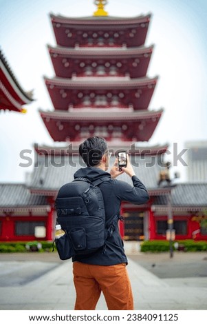 Back view of Hispanic anonymous man traveler with backpack taking photos with smartphone on street near decorated Japanese Sensoji temple Asakusa during trip in Tokyo, Japan