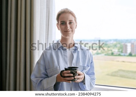 Young calm dreaming woman with cup in her hands, home window background