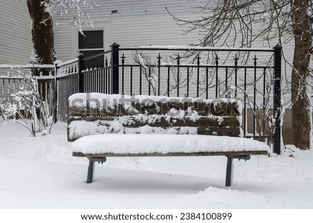 A wooden park bench on a street corner with a wrought iron fence and white wooden building in the background. The seat is covered in fresh white fluffy snow and ice. The ground is snow covered. 