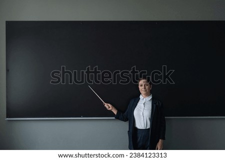 Red-haired caucasian woman in a trouser suit. Smiling female teacher with a pointer at the blackboard.  Royalty-Free Stock Photo #2384123313