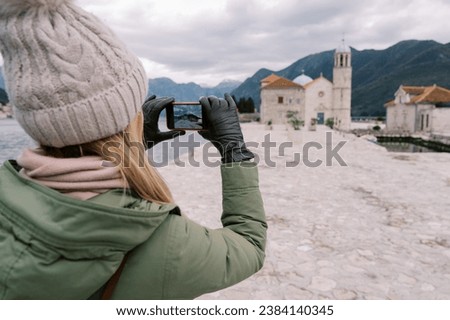 Young woman takes a picture of the Church of Our Lady on the Rocks with her smartphone. Back view. Montenegro