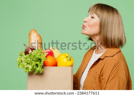 Side profile view elderly woman wear brown shirt casual clothes hold pov send blow air kiss kiss shopping paper bag with food products isolated on plain green background. Delivery service from shop Royalty-Free Stock Photo #2384177611