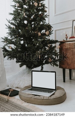 Laptop computer with blank screen on pillow, decorated Christmas tree. Bright cozy elegant home office working space interior design. Aesthetic work at home concept. Blog, website branding template