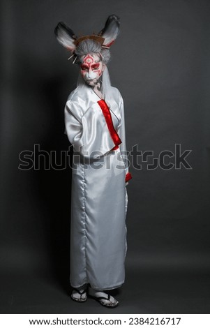 Portrait of Actress lady with stage makeup wearing rabbit mask and asian costume. Model in white kimono on black background. Halloween, carnival, performance and theater concept