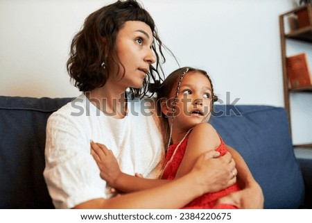 Side view portrait of young mother supporting her child, hugging frightened little daughter on sofa scared of lightning and thunder outside looking aside with anxious facial expression Royalty-Free Stock Photo #2384220675