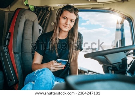 Portrait of beautiful young woman professional truck driver sitting and resting after long drive. She is using her smart phone for communication. Inside of vehicle. People and transportation concept.