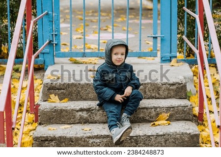 A small lonely sad child, a boy, sits on the steps in the fall near a closed kindergarten on vacation. Photography, portrait, childhood concept.