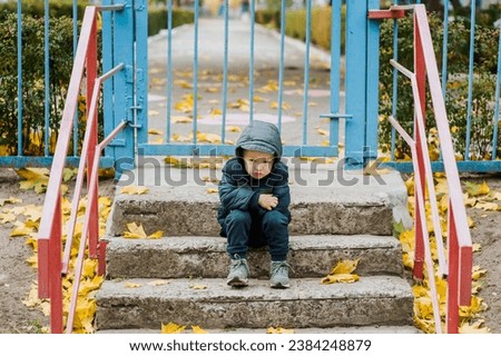 A small lonely sad child, a preschool boy, sits on the steps in the fall near a closed kindergarten on vacation. Photography, portrait, childhood concept.