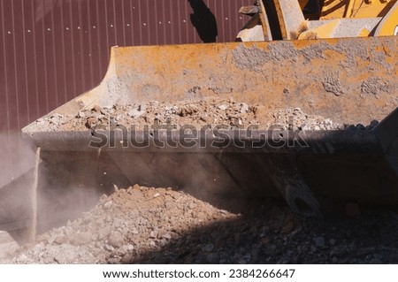 Close-up of a wheel loader bucket with rocky soil. The work of special equipment on a construction site. Sunny day. No people. Dry dusty mixture of sand and stones. Royalty-Free Stock Photo #2384266647